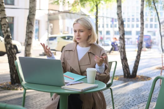Portrait of young blond woman, modern female model sitting in outdoor cafe, drinking coffee, connects to online video chat via laptop, talking to coworkers remotely, gesturing and explaining smth.