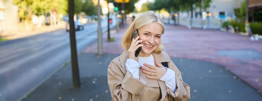Portrait of cheerful, young candid woman walking on street, answers phone call, talks on smartphone and standing near road, smiling and looking happy. Lifestyle and people concept