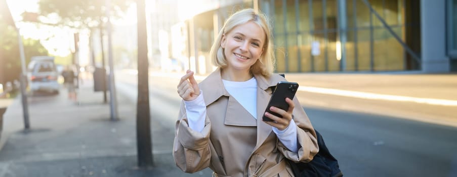 Portrait of cute blonde woman, smiling, standing on street with mobile phone, showing finger heart gesture, posing in trench coat with backpack.