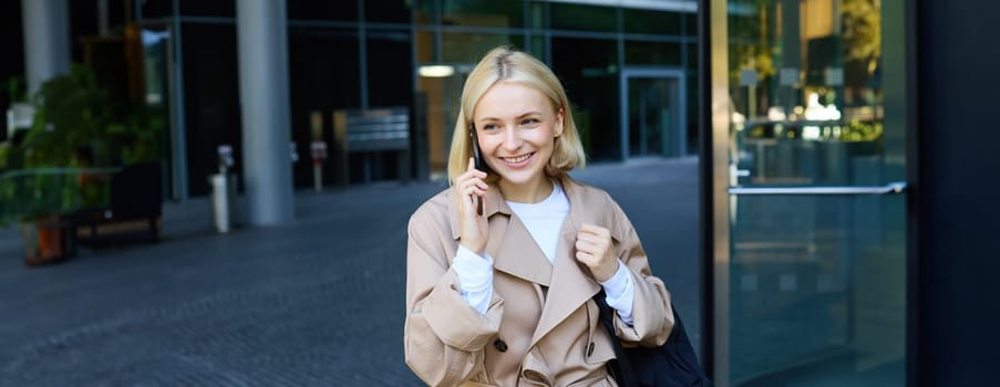 Carefree young woman with smartphone, walking outdoors in city centre, standing on street and chatting, talking on mobile phone with happy, cheerful smiling face expression.
