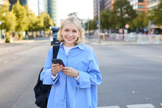 Portrait of blond smiling woman in blue shirt, holding backpack and smartphone, waiting for her ride, order taxi and checking updates on mobile phone, standing on street near road.