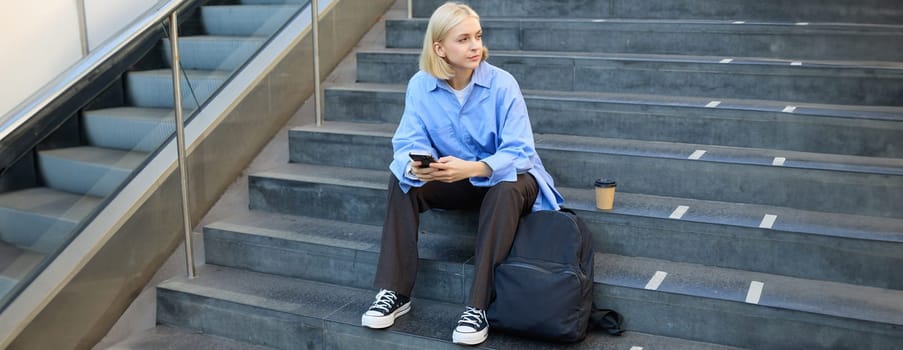 Portrait of young modern woman, student with backpack, sitting on city stairs on lunch break, using smartphone, looking aside.