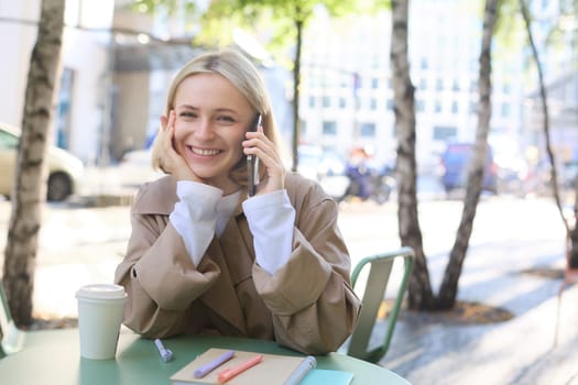 Portrait of young beautiful woman in cafe, sitting outdoors on bright sunny day, talking on smartphone, chatting over the phone and smiling.