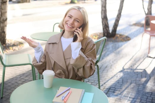 Outgoing young smiling woman, sitting in outdoor cafe, drinking coffee, answer phone call, talking on mobile, chatting with friendly attitude on smartphone.