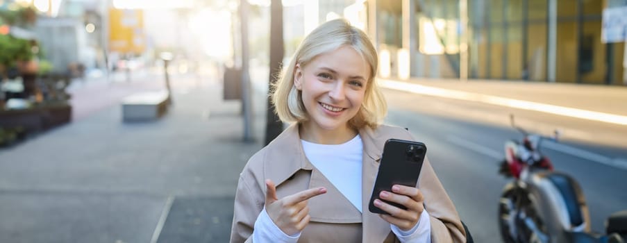Close up portrait of young blond woman, student pointing at smartphone, standing on street and showing smth on mobile phone.