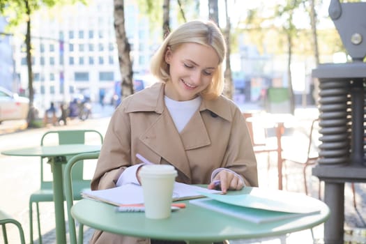 Beautiful blond woman in cafe, drinking coffee from takeaway cup, writing in notebook, checking documents on lunch break outdoors.