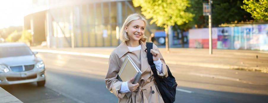Image of young modern woman, student with backpack and notebooks, female model on street, going to workplace, wearing stylish trench on sunny autumn day.