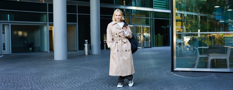 Image of stylish, modern young woman on street, posing in trench coat with backpack, standing and smiling, holding smartphone.