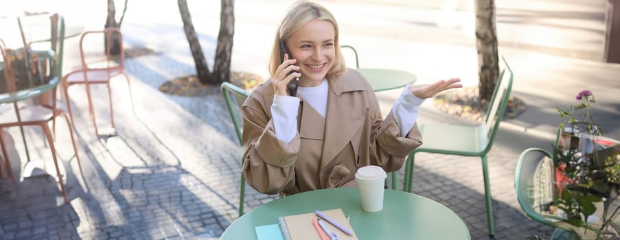 Close up portrait of happy, charismatic young woman talking on mobile phone, sitting in cafe alone, outside on sunny day, answer a call, having friendly conversation.