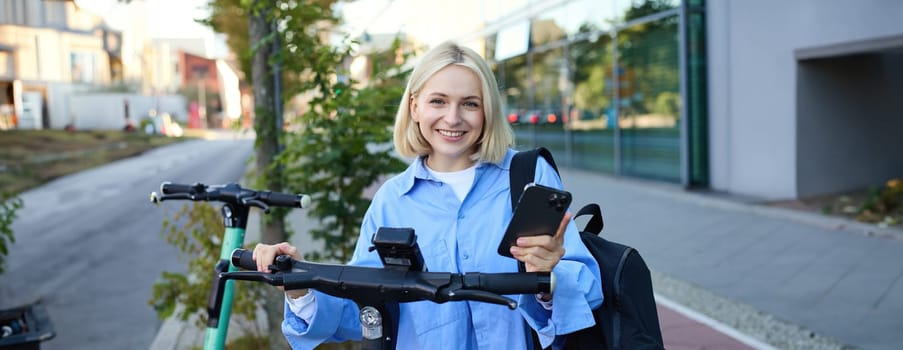 Portrait of young smiling woman, unlocks electric scooter, scans qr code with smartphone app to unlock her ride, standing on street near building.