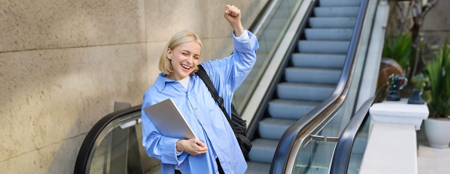 Excited girl student, woman in blue shirt, makes fist pump, stands near escalator and celebrating, feeling joy and cheer, posing with laptop and backpack. Lifestyle concept
