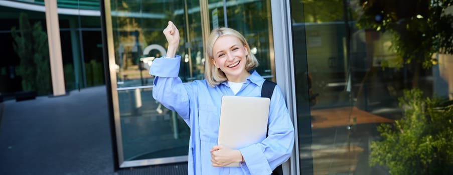 Portrait of happy, enthusiastic blond woman near office building, holding backpack and laptop, raising hands up and cheering, encouraging you, chanting and smiling.