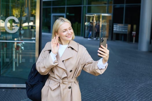 Lifestyle portrait of young modern woman, cute girl video chatting on street, waving hand at her smartphone and smiling, walking on street and talking to friend on mobile phone app.