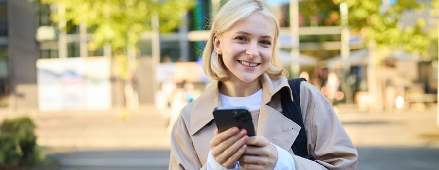 Close up portrait of smiling, beautiful blond girl, holding backpack and mobile phone, standing on street with lots of sunlight, buildings and road behind, looking happy at camera.