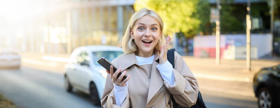 Portrait of amazed blonde female model on street, standing with mobile phone and gasping in surprise, hears about amazing promo.