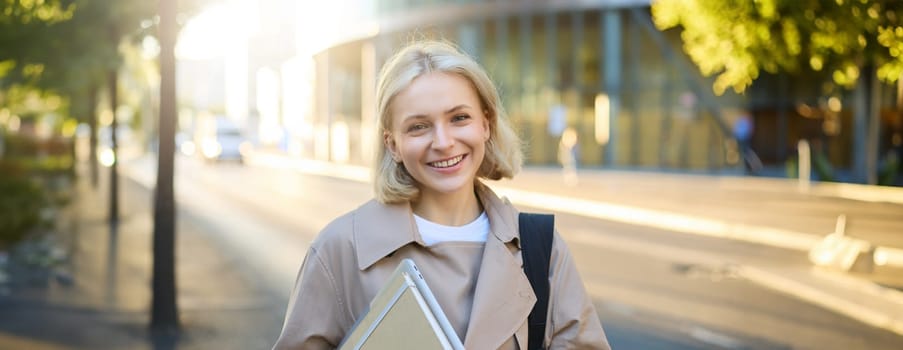 Image of beautiful young smiling woman with blond hair, walking along street on sunny day, carry her notebooks and college materials, looking carefree.