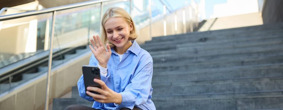 Cute smiling young woman, sitting on stairs with mobile phone, waving hand and saying hello during video chat, connects to online meeting.