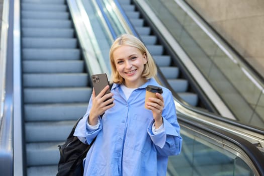 Image of stylish young woman in blue shirt, holding takeaway cup of coffee and mobile phone, standing near escalator and waiting for you, smiling at camera.