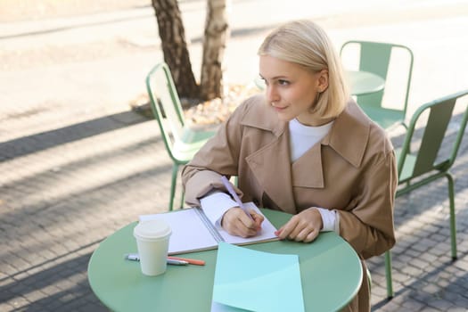 Portrait of woman studying outside, sitting in cafe with notebook, doing homework and drinking coffee, looking aside. Lifestyle and people concept