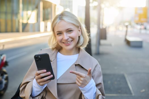 Close up portrait of young blond woman, student pointing at smartphone, standing on street and showing smth on mobile phone.