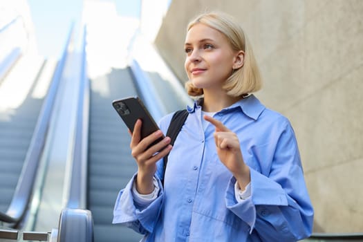 Portrait of beautiful young blond woman in blue shirt, standing near escalator, holding mobile phone, using smartphone, waiting for someone in city centre.
