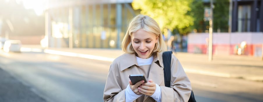Portrait of surprised blonde woman, standing on street, looking at her mobile phone with amazed face, reading great news on smartphone.