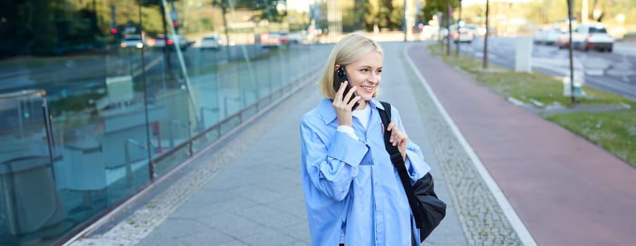 Cheerful young modern woman walking on street and answers phone call, talking on mobile, carries black backpack.