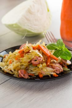 cooked fried cabbage with vegetables and sausages, in a plate on a wooden table.