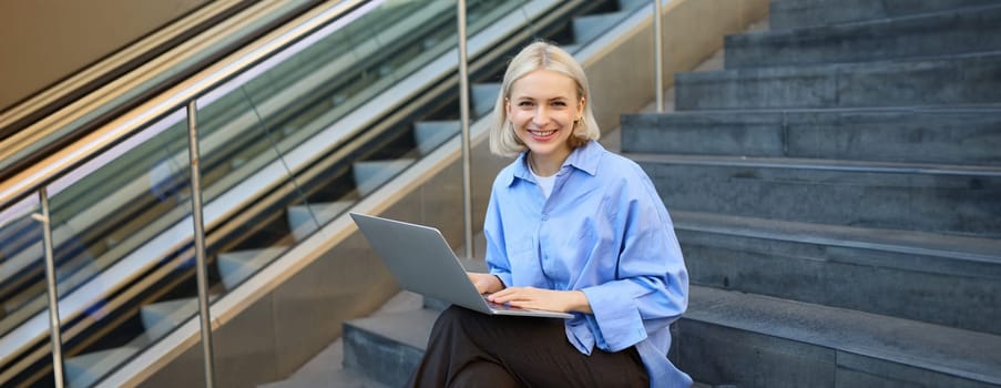 Girl student sitting on stairs in university campus, using laptop, working on project, doing homework, e-learning outdoors, drinking coffee outside, wearing stylish blue shirt.