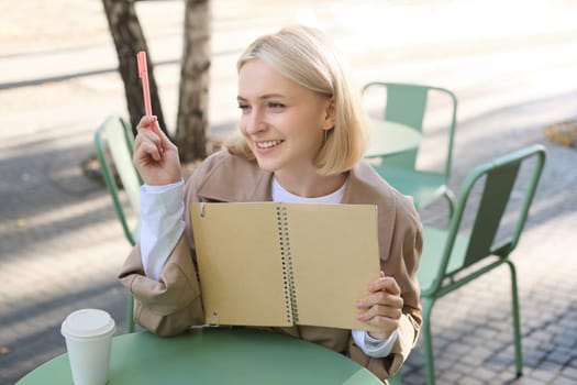 Portrait of young beautiful blond woman, artsy girl in coffee shop, holding notebook and pen, writing in her journal, drawing sketches outdoors.