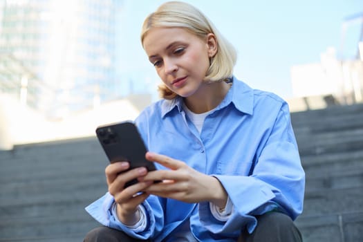 Close up portrait of young stylish female model, sitting outdoors on stairs, using mobile phone, reading on smartphone, messaging while waiting for someone outside.