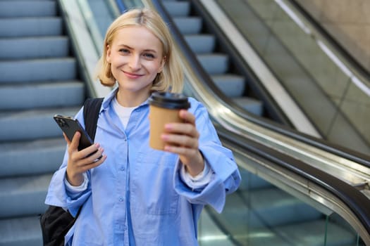 Portrait of smiling, beautiful young woman with smartphone, standing on bottom of escalator, giving you cup of coffee, brought takeaway.