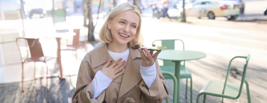 Image of beautiful female model, smiling, recording voice message, translating something using smartphone app and looking happy, sitting outdoors in cafe.