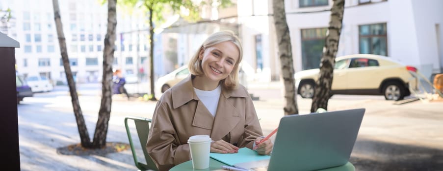 Remote workplace. Smiling young woman freelancer, student using laptop in outdoor cafe, talking to someone, attends online lecture, drinking coffee.