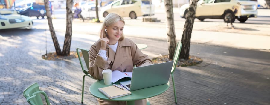 Young woman, freelancer, student doing homework in outdoor cafe, drinking her coffee on street, using laptop, connects to online lecture or doing course in internet.