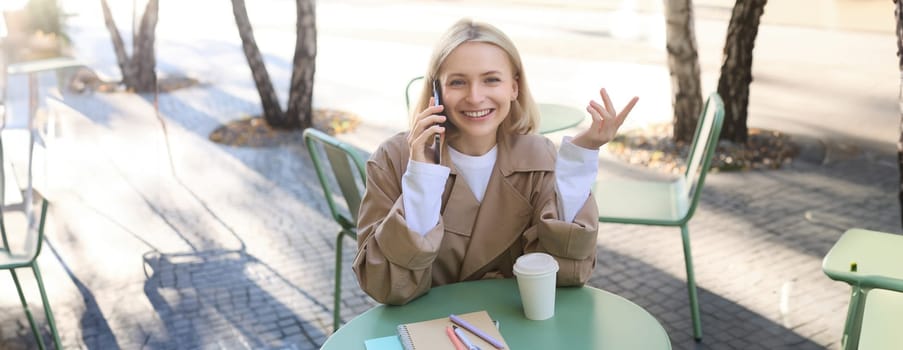 Image of stylish blond woman talking on mobile phone, drinking coffee in cafe outdoors, enjoying warm weather in city centre, answer telephone call on smartphone.