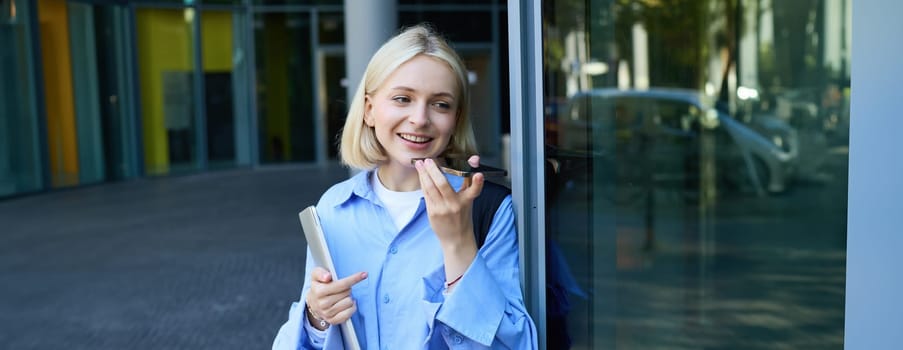 Portrait of beautiful, smiling young student, woman records voice message, talking on speakerphone, standing near office building on street and holding laptop with backpack.