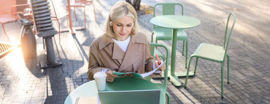 Portrait of young blond woman, female student in street cafe, wearing wireless headphones, using laptop, having online meeting, attend web lecture or course, doing homework.
