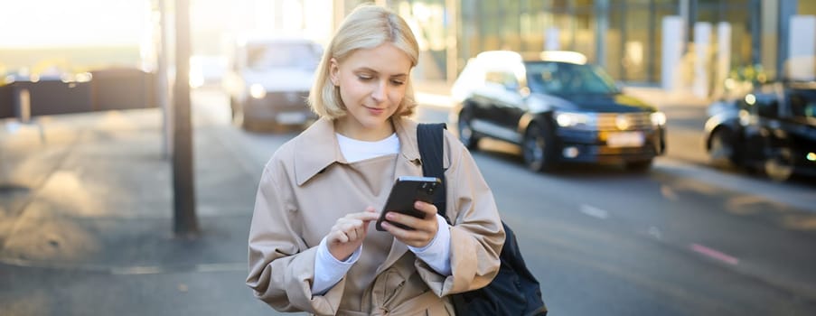 Close up portrait of young modern woman on street, using mobile phone, looking at smartphone screen, reading notification, waiting for taxi near road.