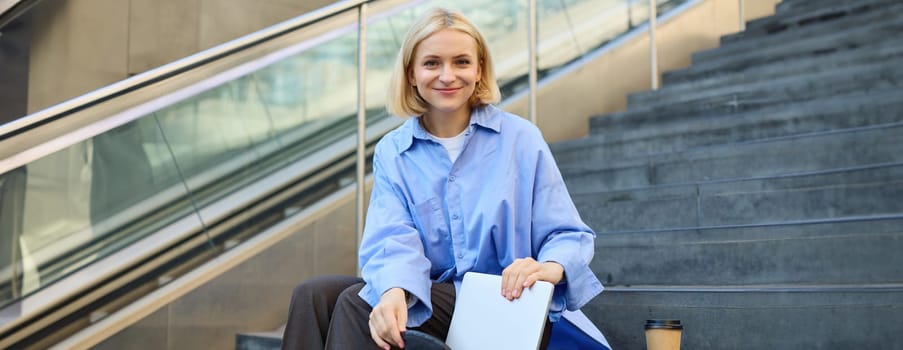 Portrait of beautiful smiling woman, student on stairs in city, sitting and drinking coffee, packing laptop in backpack, looking happy at camera.