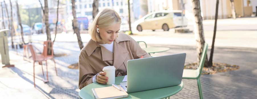 Portrait of young blond woman with laptop, sitting on street, drinking coffee in outdoor cafe, working, wearing wireless headphones, doing homework, freelancing.