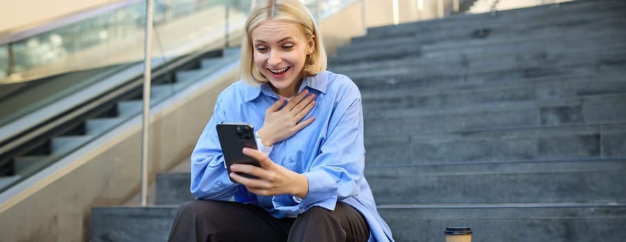 Portrait of young blond woman, sitting outside campus on city stairs, looking at smartphone with happy surprised face, receive positive good news on video chat, watching interesting video.