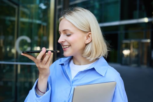 Close up portrait of smiling young female student, woman records voice message on smartphone, speaking on speakerphone while standing near office building, holding laptop.