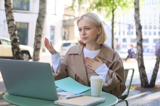 Image of woman talking, introducing herself during online meeting, using laptop in outdoor cafe, working remotely, drinking coffee.