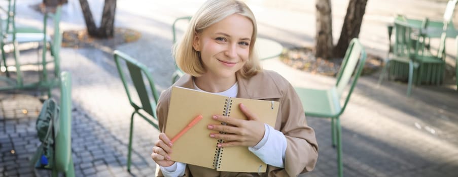 Outdoor shot of young beautiful blond girl with notebook, woman sitting in outdoor cafe, writing in journal, making plans in her planner, putting notes.