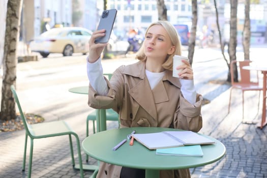 Fashionable young blond woman, taking selfie with favourite drink in coffee shop, puckers lips, creating content for social media, influencer making photos.
