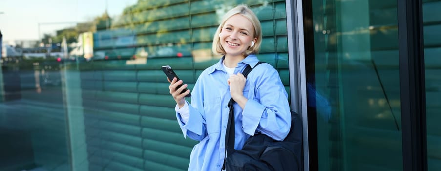 Lifestyle portrait of smiling young female model, student with backpack, waiting for someone on street, standing outdoors with smartphone.