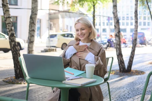Image of young smiling woman working remotely, connects to online call, chatting via laptop, looking pleased, feeling grateful, laughing, sitting in outdoor cafe.