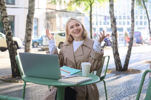 Image of young modern woman, chatting with someone online, having video call via laptop, sitting in outdoor cafe, drinking coffee, gesturing and smiling.