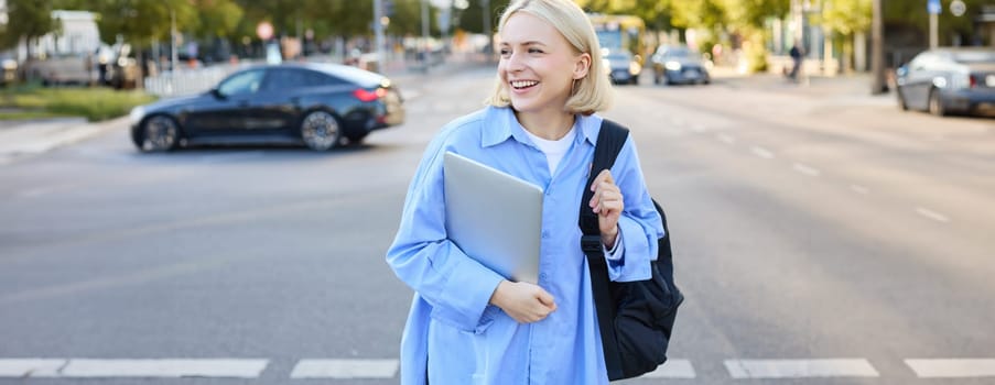 Lifestyle portrait of candid young woman, walking on street, laughing and smiling, holding backpack and laptop, looking aside with happy smiling face.